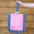 A hand holding a book bag with a pink and blue checkered pattern and yellow ruffles, against a wooden background.