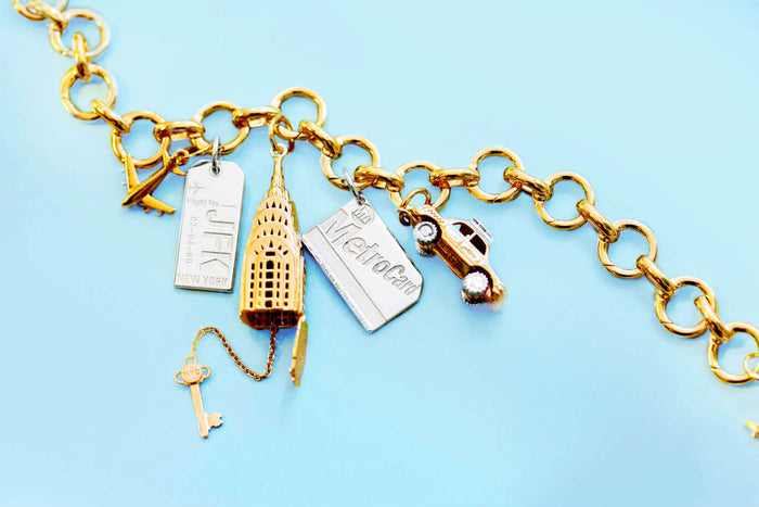 Gold charm bracelet with multiple NYC charms: silver JFK luggage tag, gold Chrysler Building with 'NYC' key, silver MetroCard, and gold taxi cab with silver taxi topper and silver wheels.