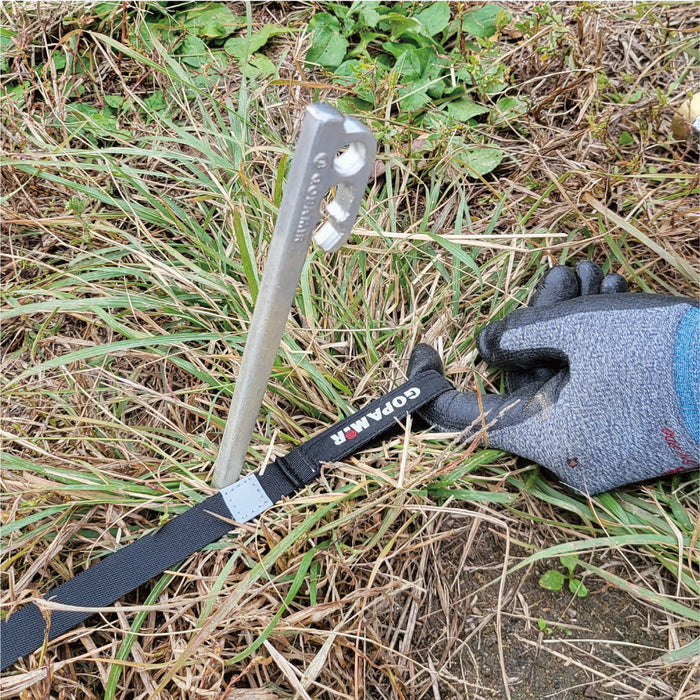 Gloved hand tensioning black Gopamir G450 guy-line webbing anchored to a silver tent stake in grass