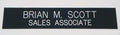 A black rectangular name tape or patch with white text reading 'BRIAN M. SCOTT SALES ASSOCIATE' on a light gray background.